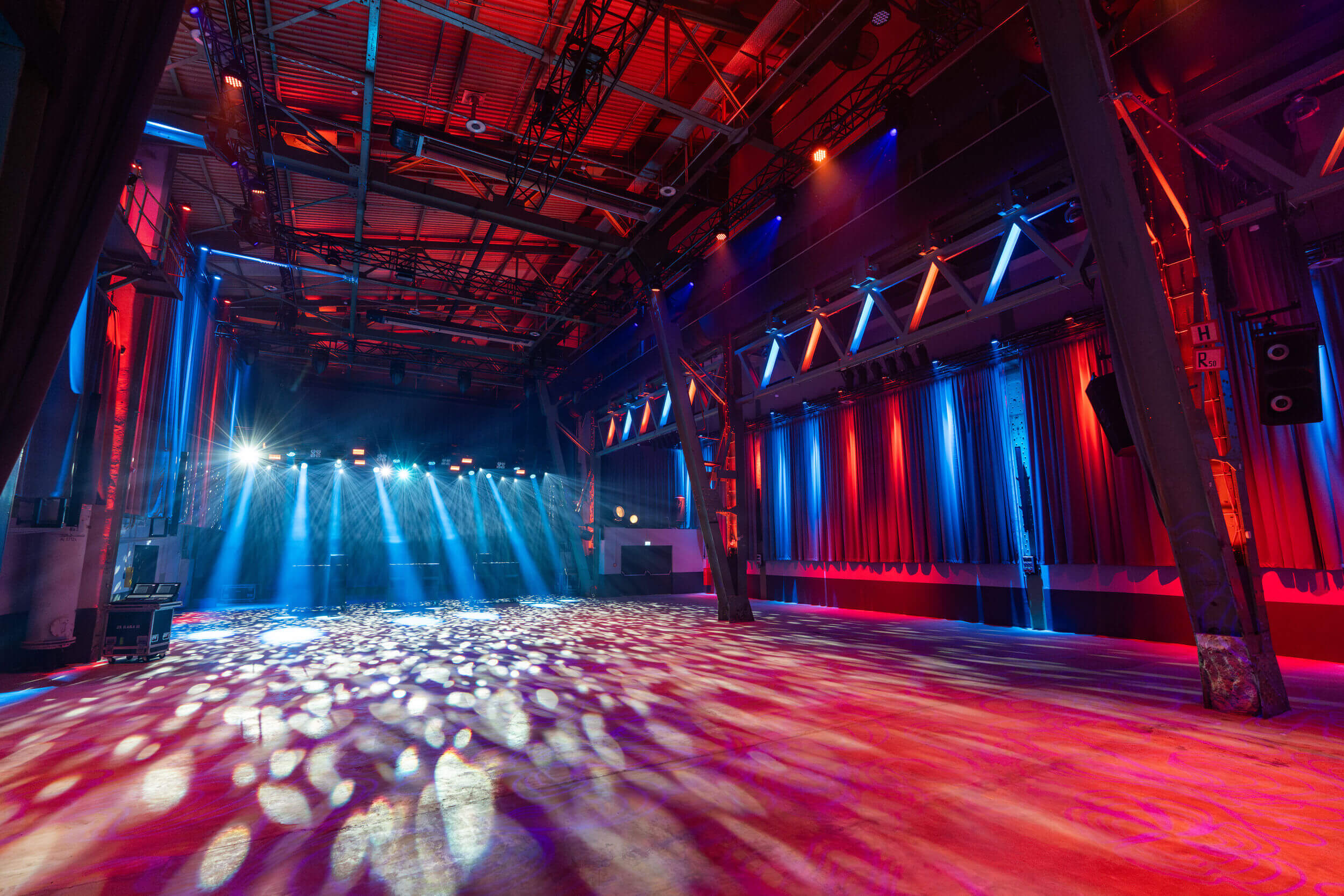 Großer leerer Veranstaltungsraum mit roter und blauer Beleuchtung; Lichtstrahlenmuster auf dem Boden und sichtbare Stahlkonstruktion.