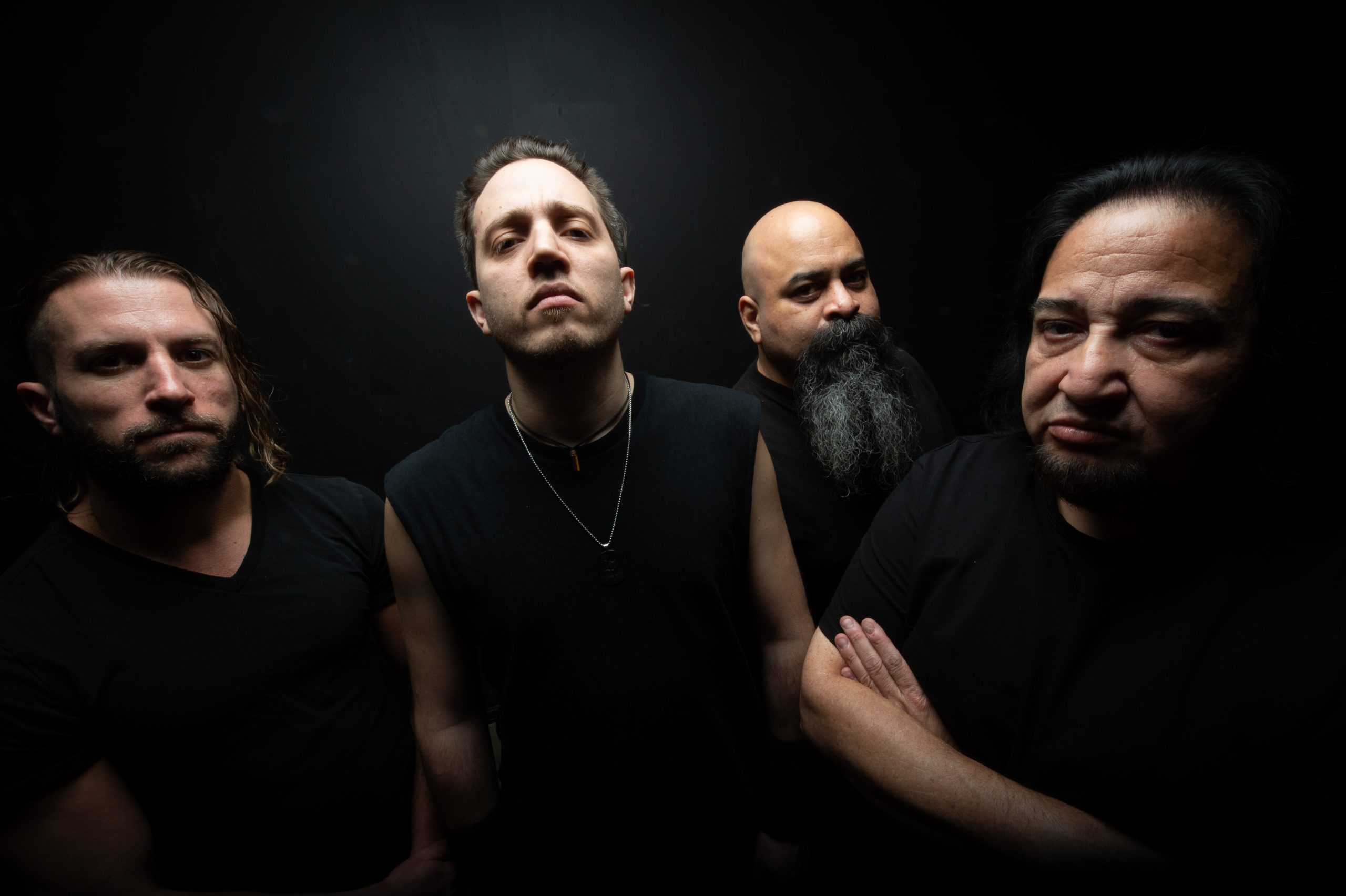Four men in black clothing pose against a dark background; the central man with short hair looks seriously ahead, the man in the back right with dark hair looks to the side, the second from the left wears a chain, and the man in the middle right has a long beard.