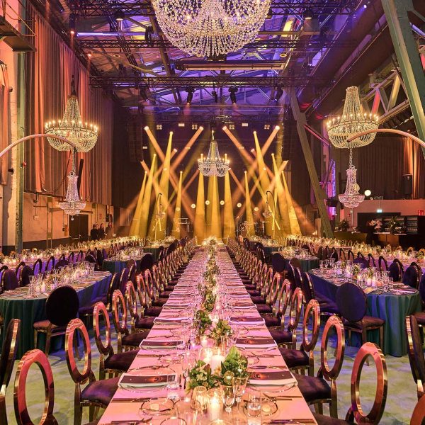 Große Festhalle mit langen Tafeln, Kerzen, Blumengestecken, eleganten Kronleuchtern und buntem Bühnenlicht im Hintergrund.
