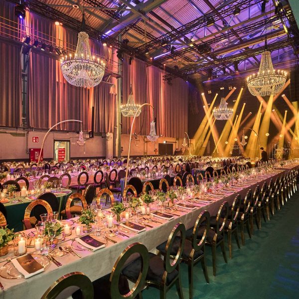 Große Gala-Tafel in einer Industriehalle mit goldenen Stühlen, Kronleuchtern, Kerzenlicht und Blumenarrangements.